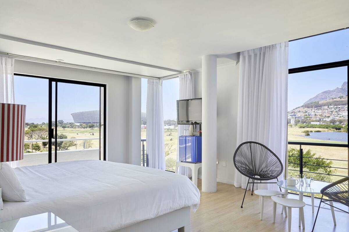 Ett modernt hotellrum med en stor säng, en TV, en balkong med utsikt över staden och bergen samt en matplats med stolar och ett bord.