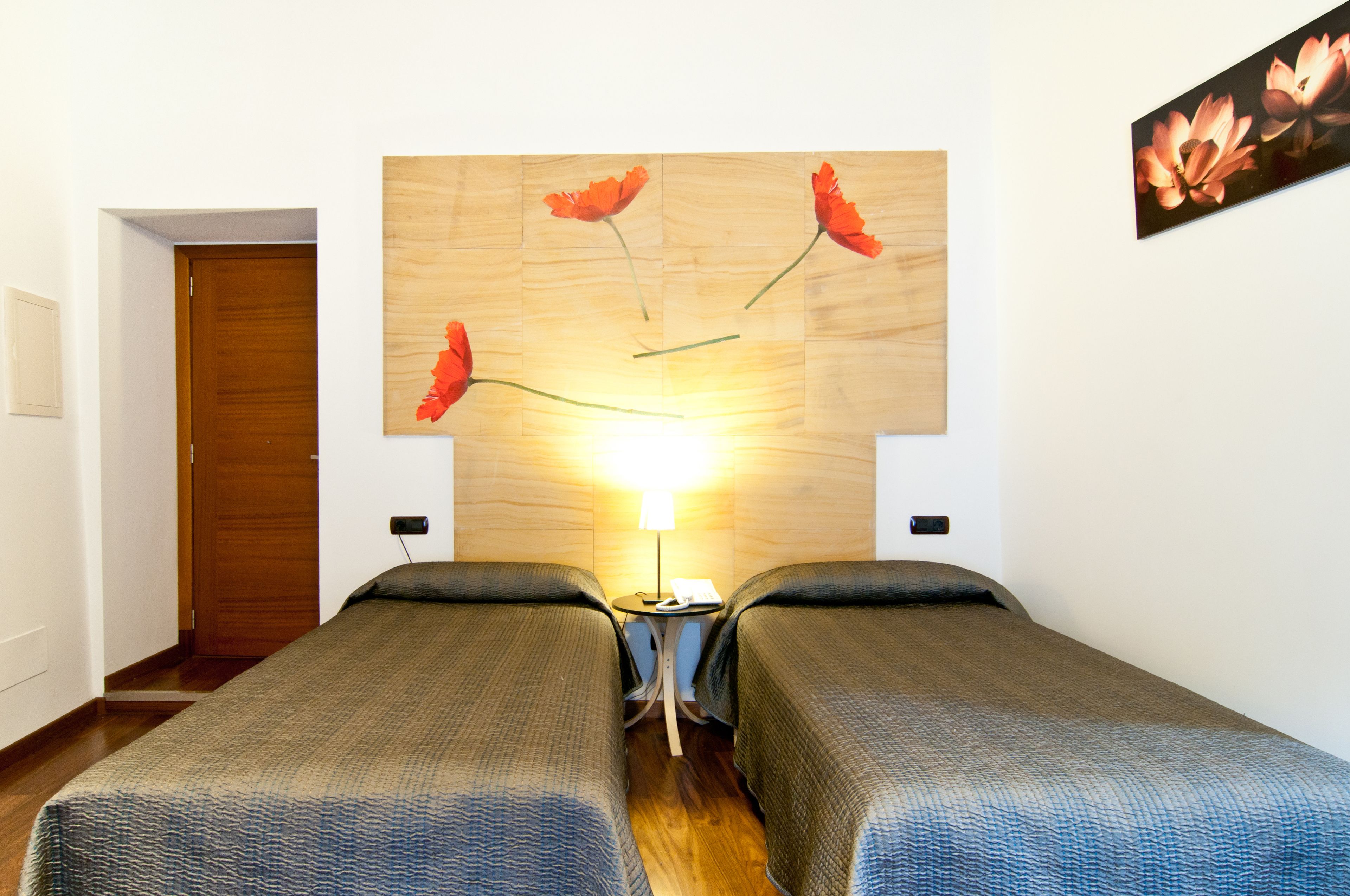 A hotel room with two single beds, a wooden headboard with a floral painting, a small table with a lamp, and a wooden door.
