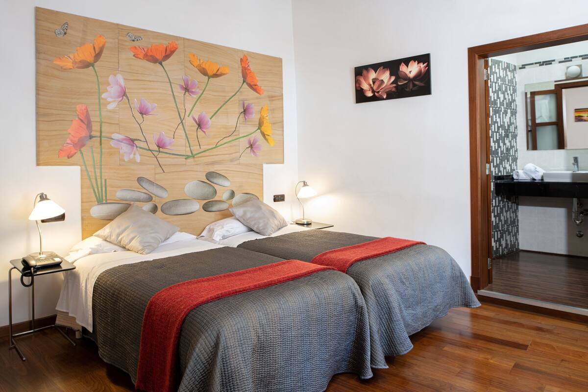 Habitación de hotel con dos camas, cabecero de madera con motivos florales, baño con espejo y lavamanos, y una mesita con lámpara.
