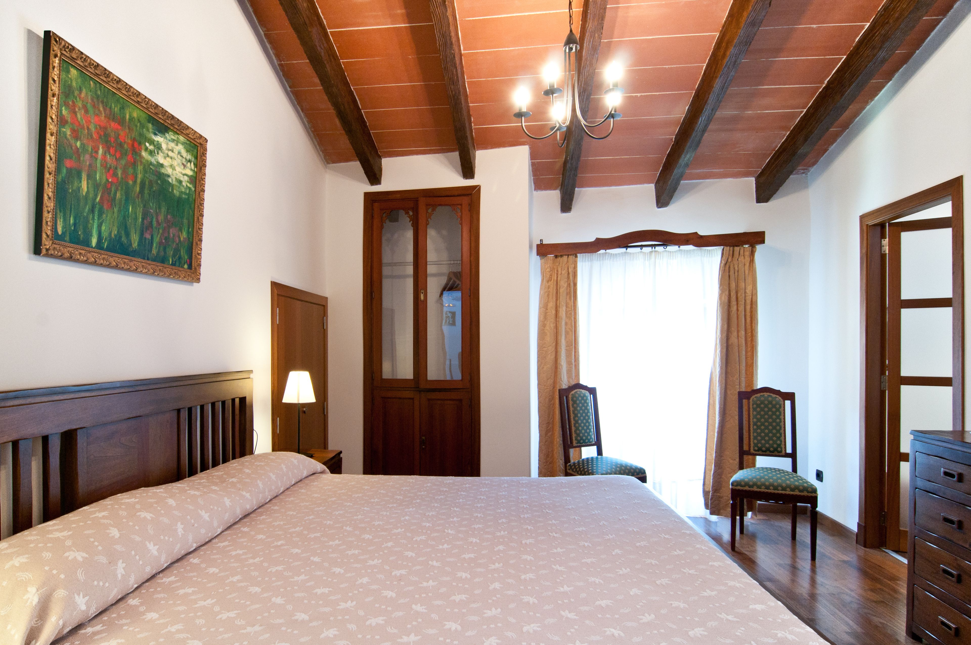 A bedroom with a large bed, wooden ceiling beams, a chandelier, a wooden door, and a window with curtains.