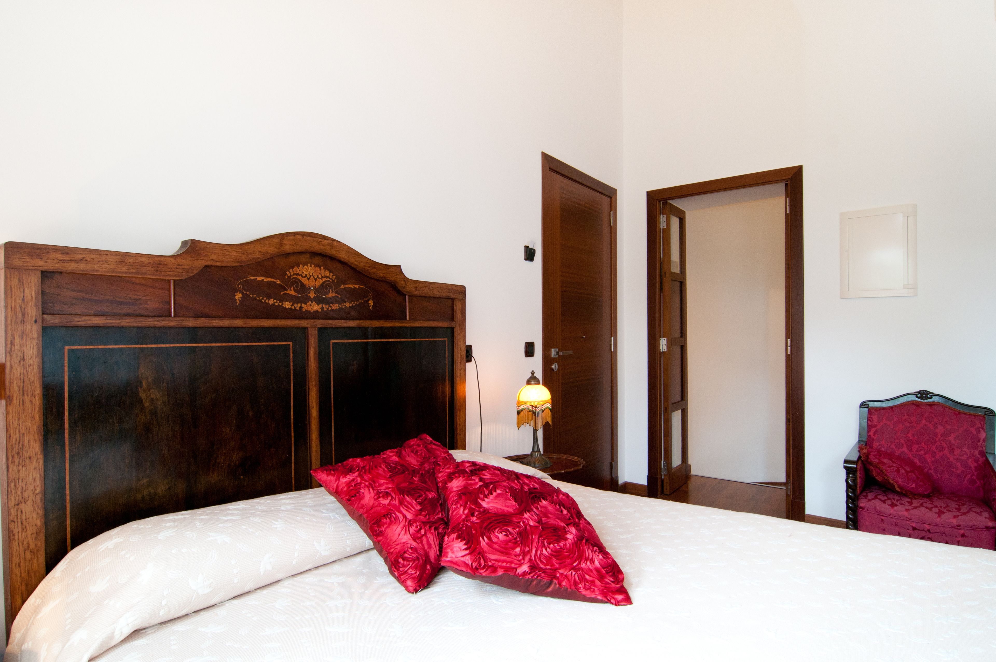 Een slaapkamer met een houten hoofdeinde, een bed met een wit en rood gebloemd sprei, een nachtkastje met een lamp en een rode stoel.