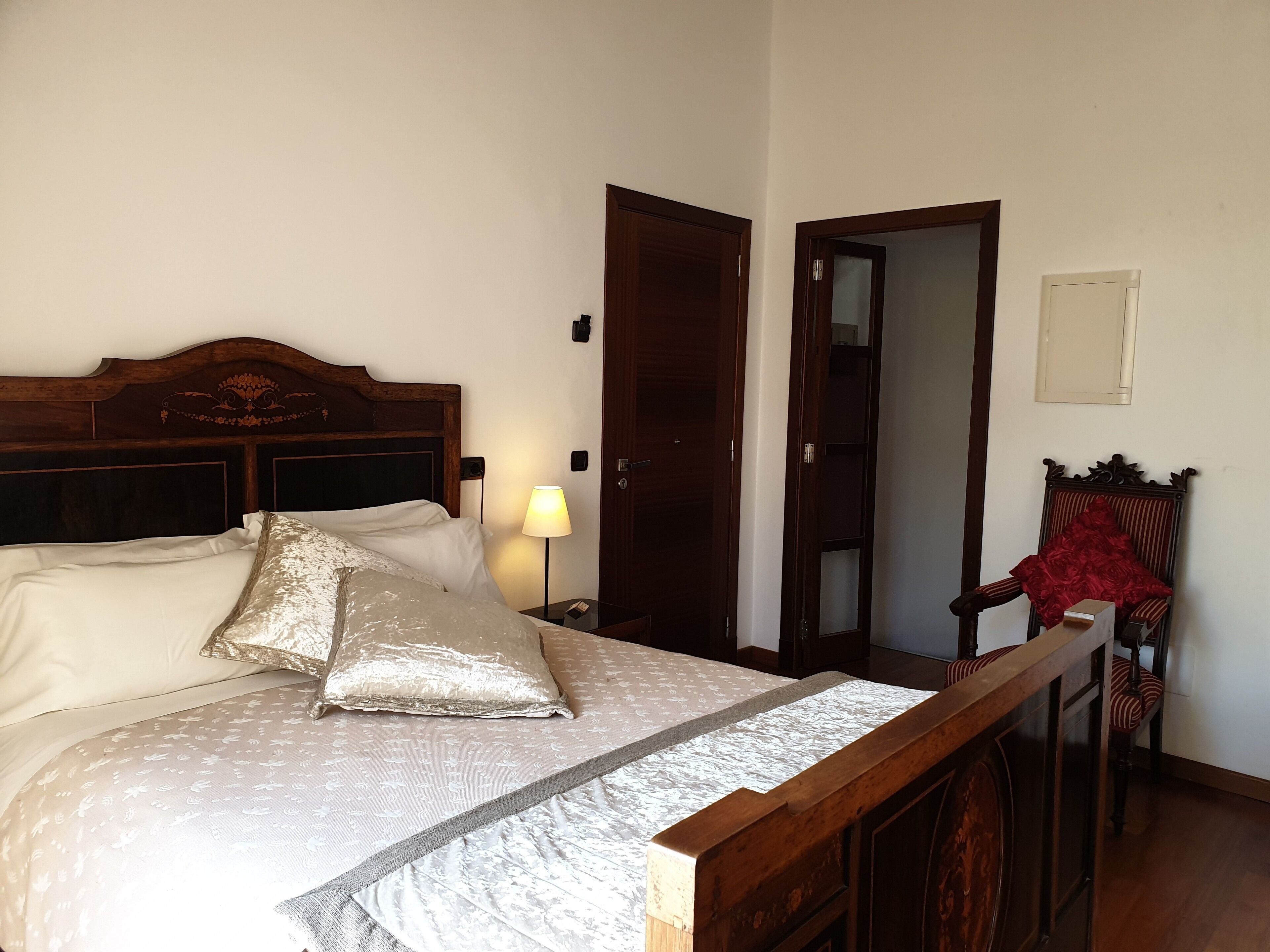 Uma cama bem arrumada com cabeceira de madeira, luminária de cabeceira e uma cadeira com capa vermelha.