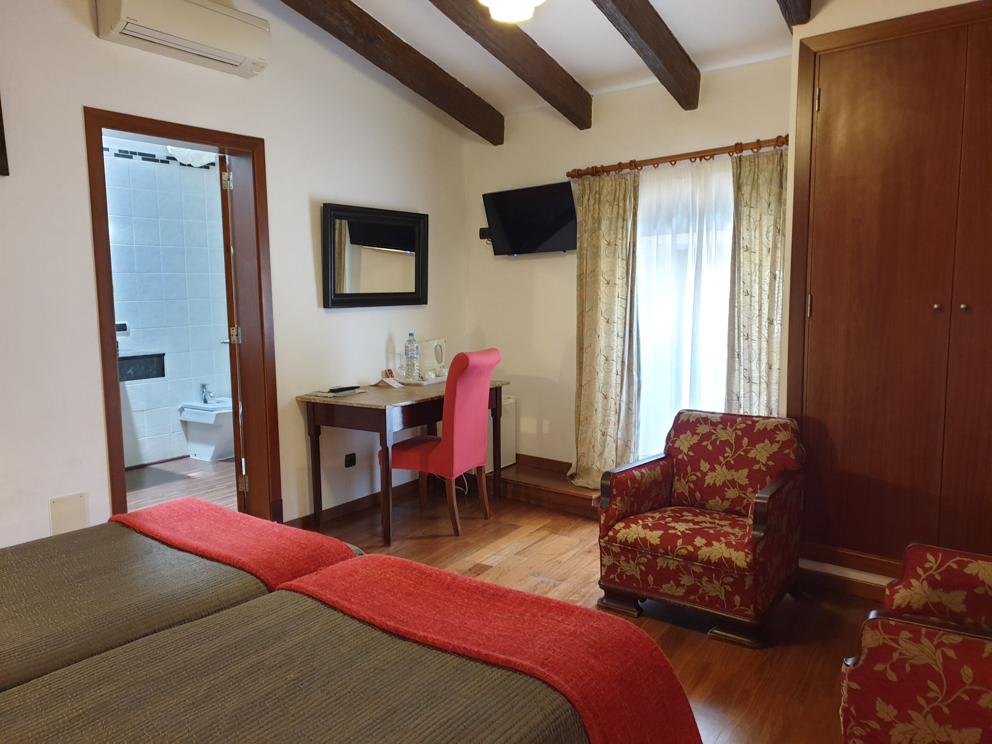 Ein Hotelzimmer mit Bett, Schreibtisch mit Stuhl, einem roten Sessel, einem Kleiderschrank und einem durch eine offene Tür einsehbaren Badezimmer.