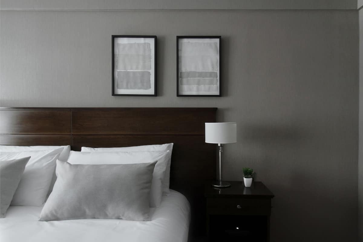 A neatly made bed with white linens and pillows, a dark wooden headboard, a bedside table with a lamp and a small potted plant, and two framed abstract artworks on the wall.