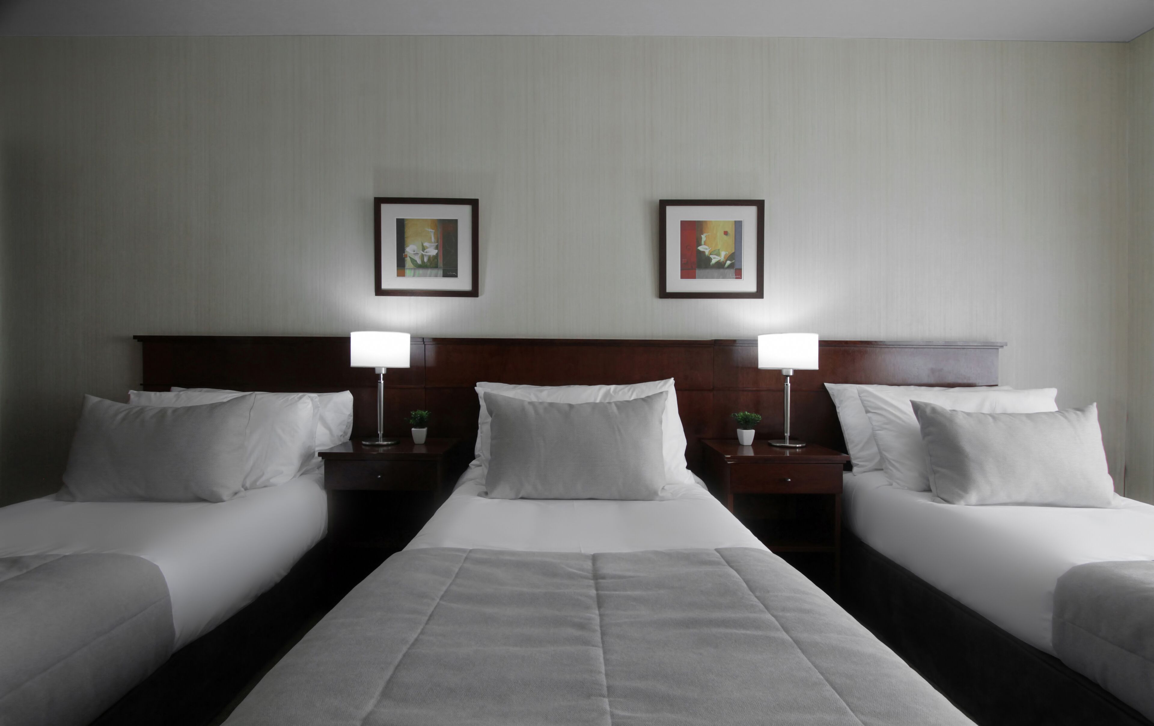A hotel room with two single beds, a large bed in the middle, and two nightstands with lamps. There are two framed pictures on the wall above the beds.