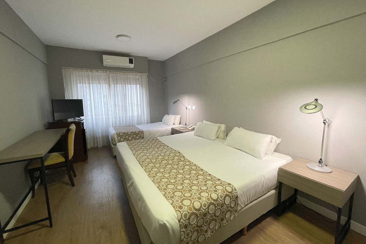A hotel room with two beds, a desk, a chair, a TV, a lamp, and an air conditioning unit.