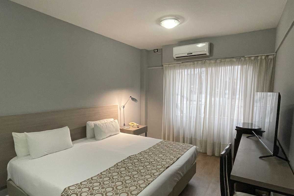 A hotel room with a large bed, bedside lamp, air conditioning unit, and a desk with a chair.