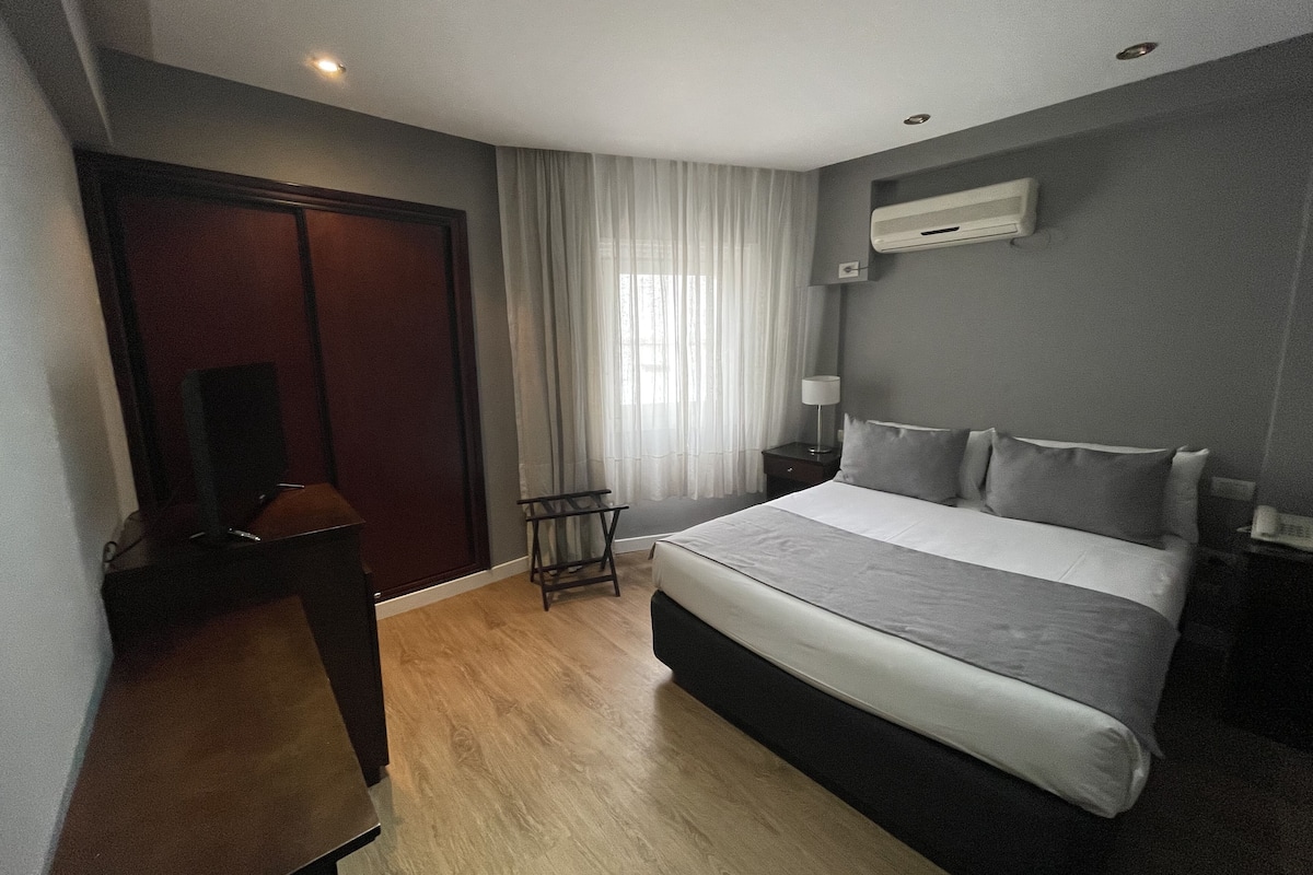 A hotel room with a large bed, a desk, a chair, a nightstand, and a window with curtains.