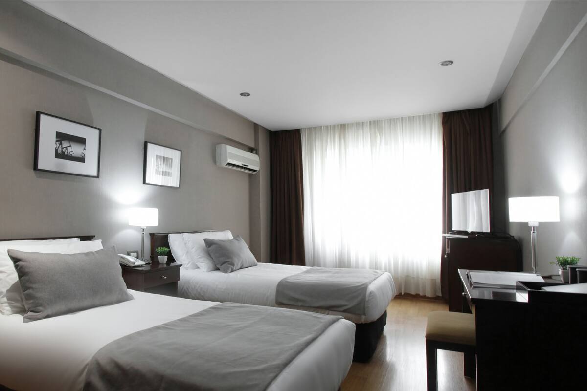 A hotel room with two beds, a desk, a chair, a lamp, and a window with curtains.
