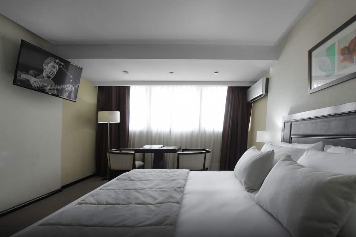 A hotel room with a large bed, a desk with two chairs, a window with curtains, and a framed picture on the wall.