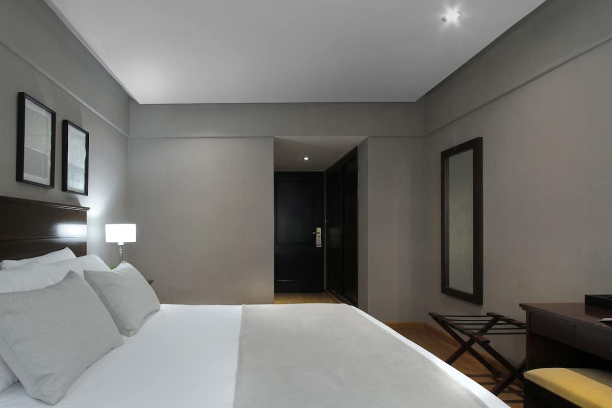A hotel room with a large bed, a desk, a chair, and a door leading to another room.