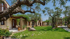 A courtyard with stone buildings, a grassy lawn, and large trees. - Paliokaliva Village (Zakynthos)