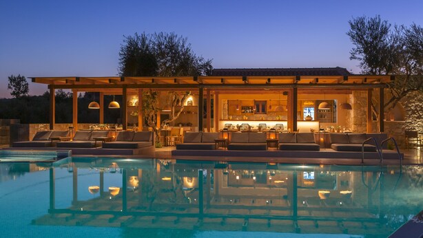 A poolside area with lounge chairs and a bar area with seating and a kitchen visible through the glass. - Paliokaliva Village (Zakynthos)