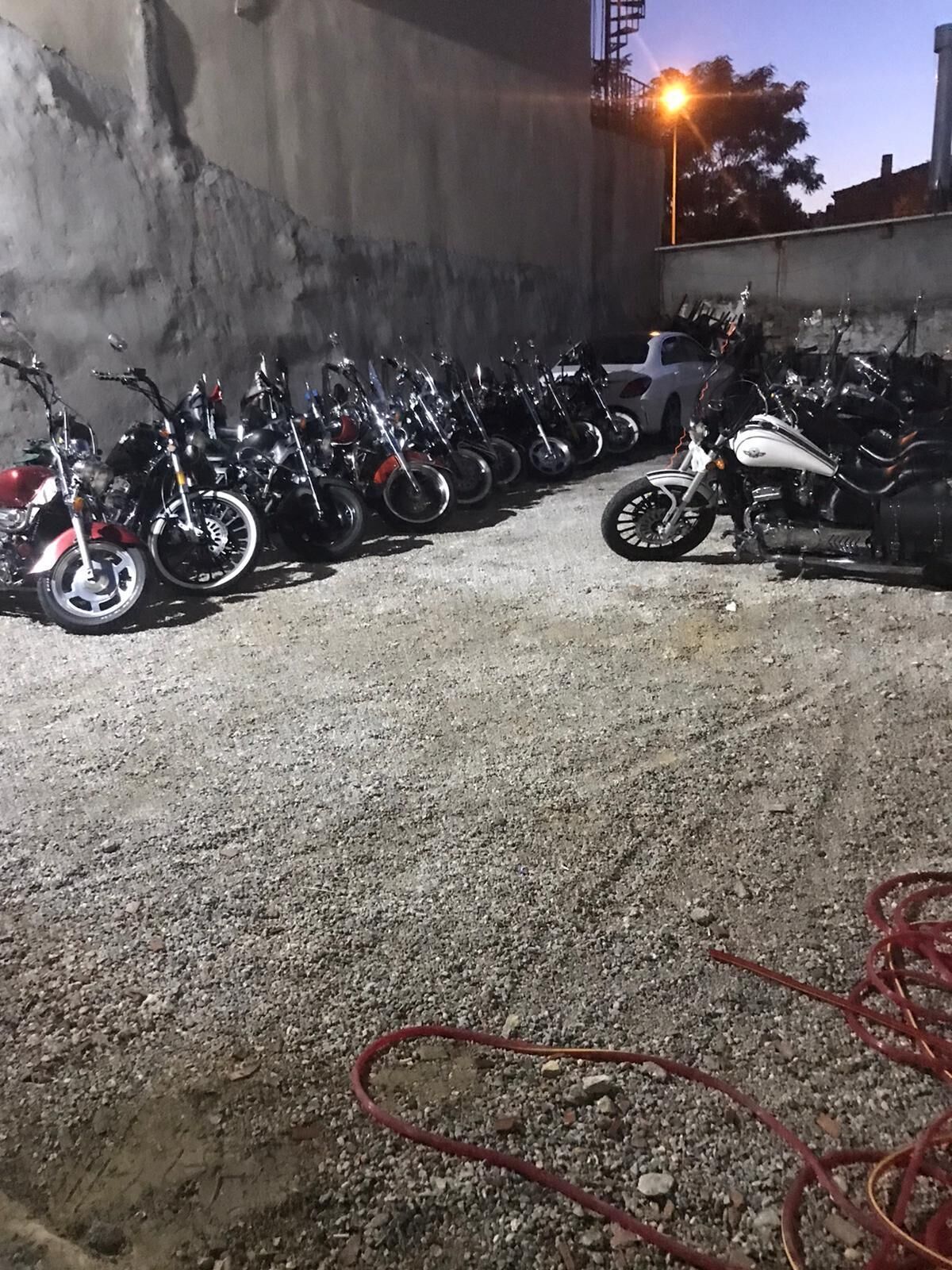 A row of motorcycles parked in a gravel area with a concrete wall in the background.