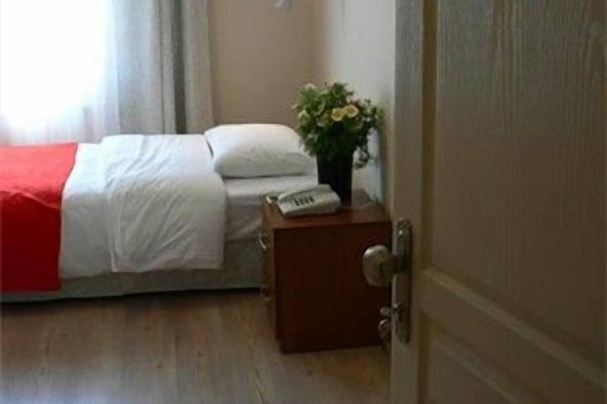 A hotel room with a bed, a bedside table with a vase of flowers, a door with the number 103, and a window with curtains.