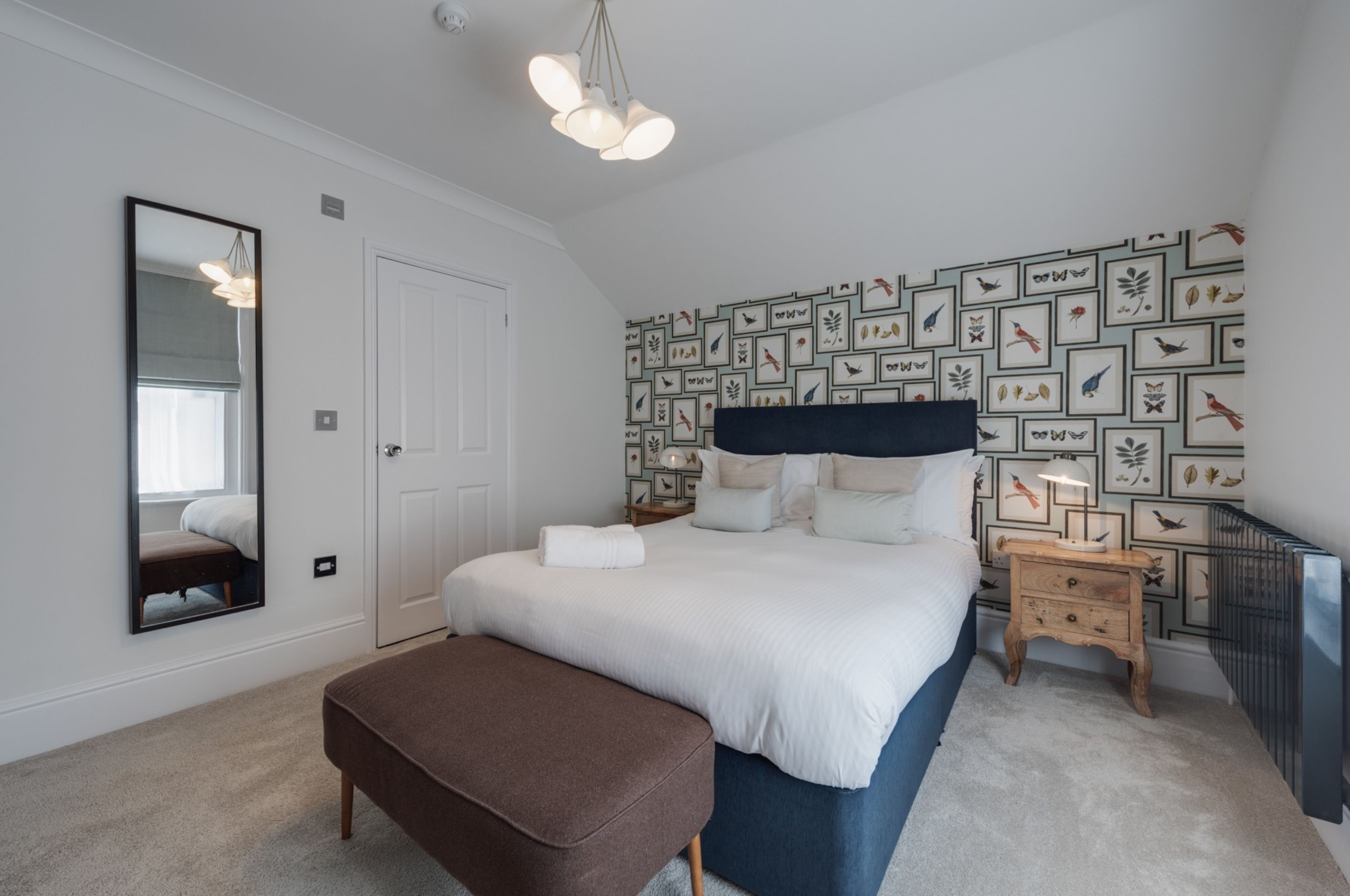 A bedroom with a large bed, a bedside table, a nightstand, a mirror, and a wall with framed pictures.