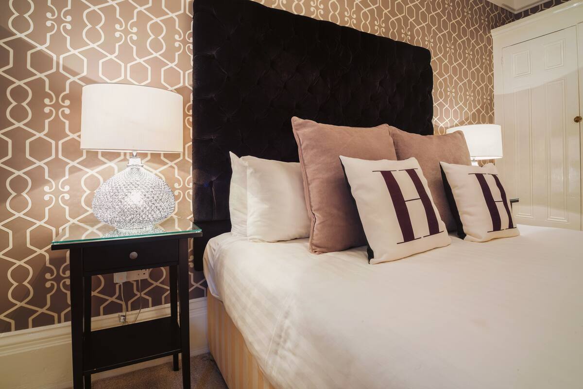Una cama bien hecha con cabecera oscura, almohadas blancas y marrones, y dos mesitas de noche con lámparas.