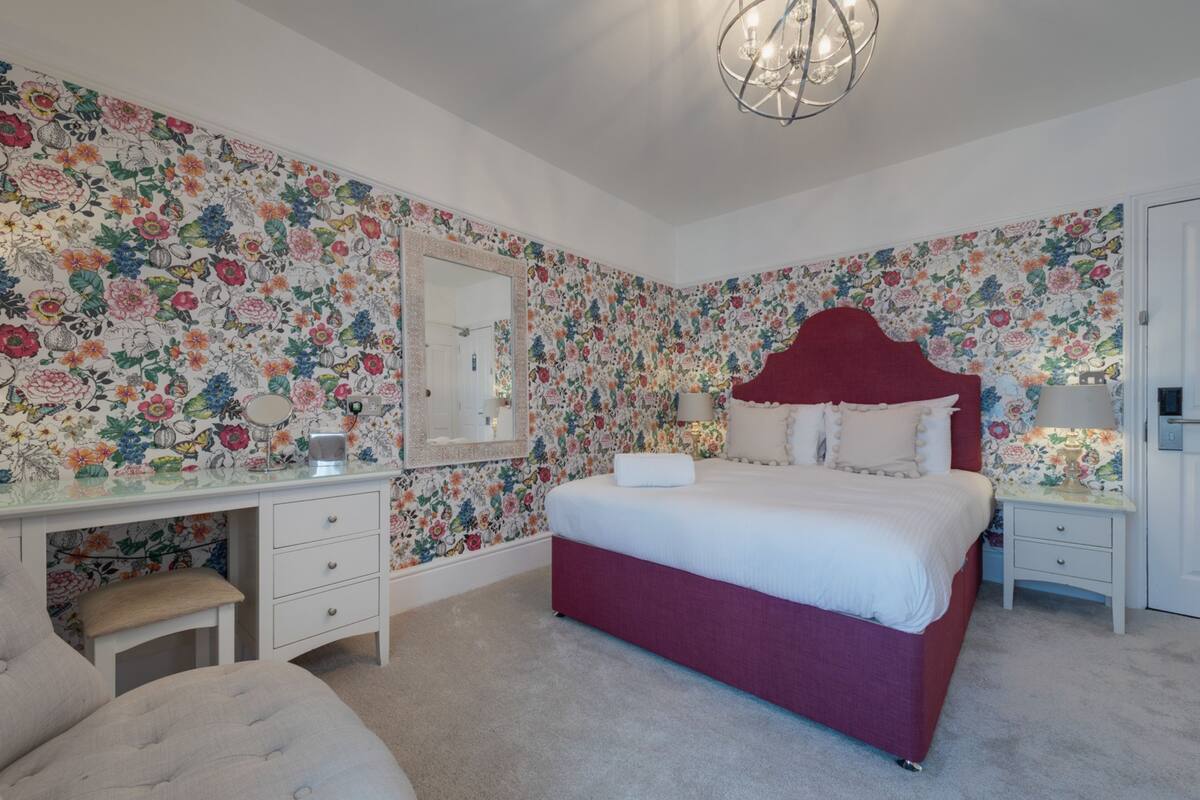 Un dormitorio con papel tapiz floral, un cabecero rojo, una cama blanca, una mesita de noche con una lámpara y un espejo.