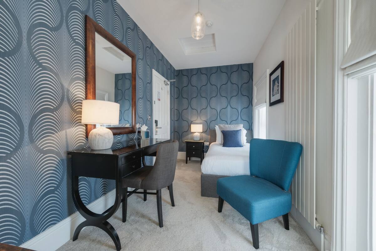 Habitación de hotel con escritorio, silla y un sillón azul. Hay un espejo grande en la pared y un cuadro enmarcado sobre la cama.