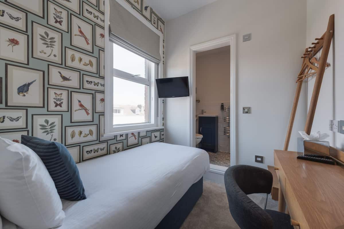 Un dormitorio con cama, escritorio, silla, televisor y baño visible a través de una puerta abierta.