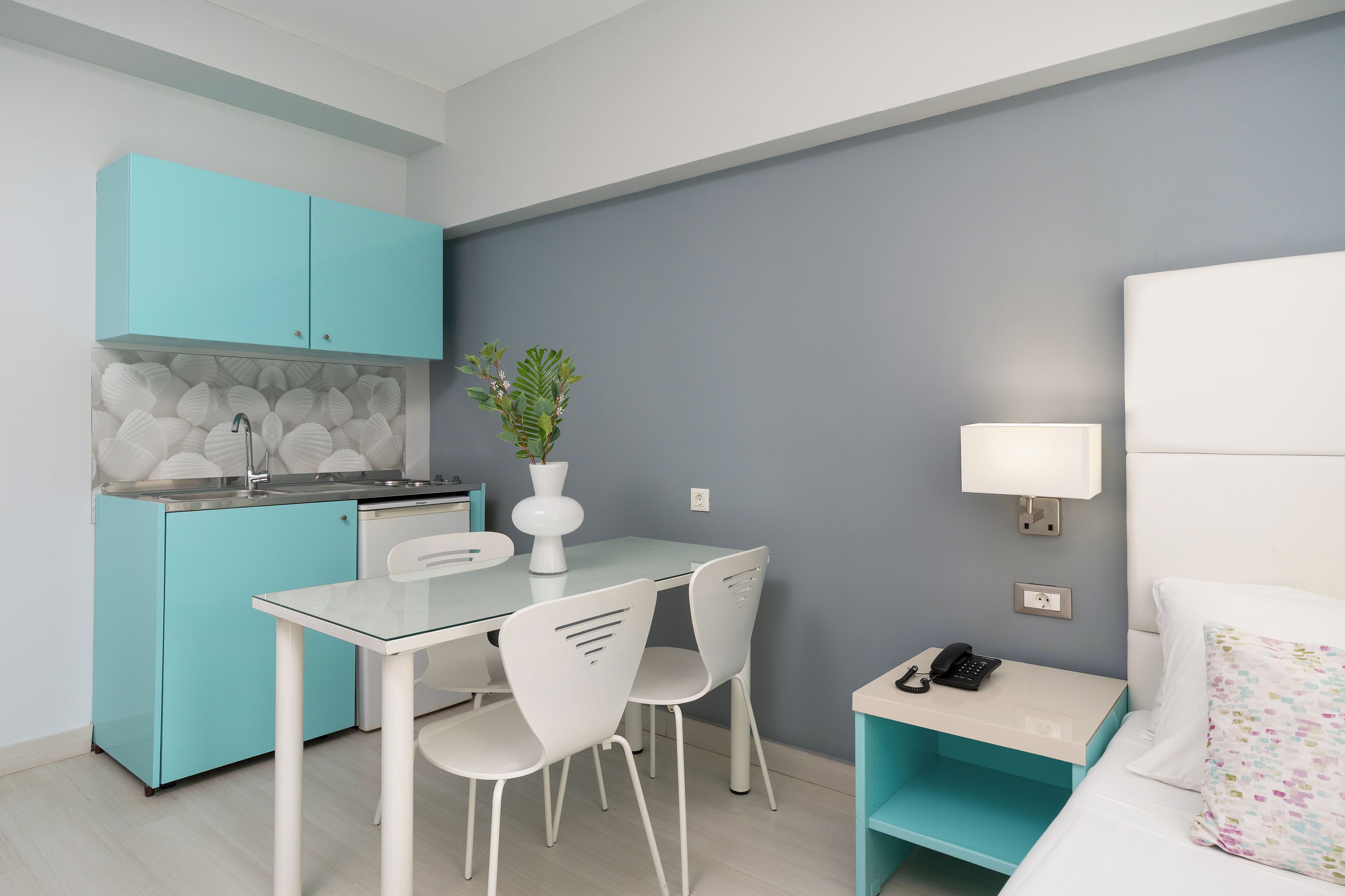 A compact room with a kitchenette, a dining area with chairs, a bed with a nightstand, a lamp, and a small plant.