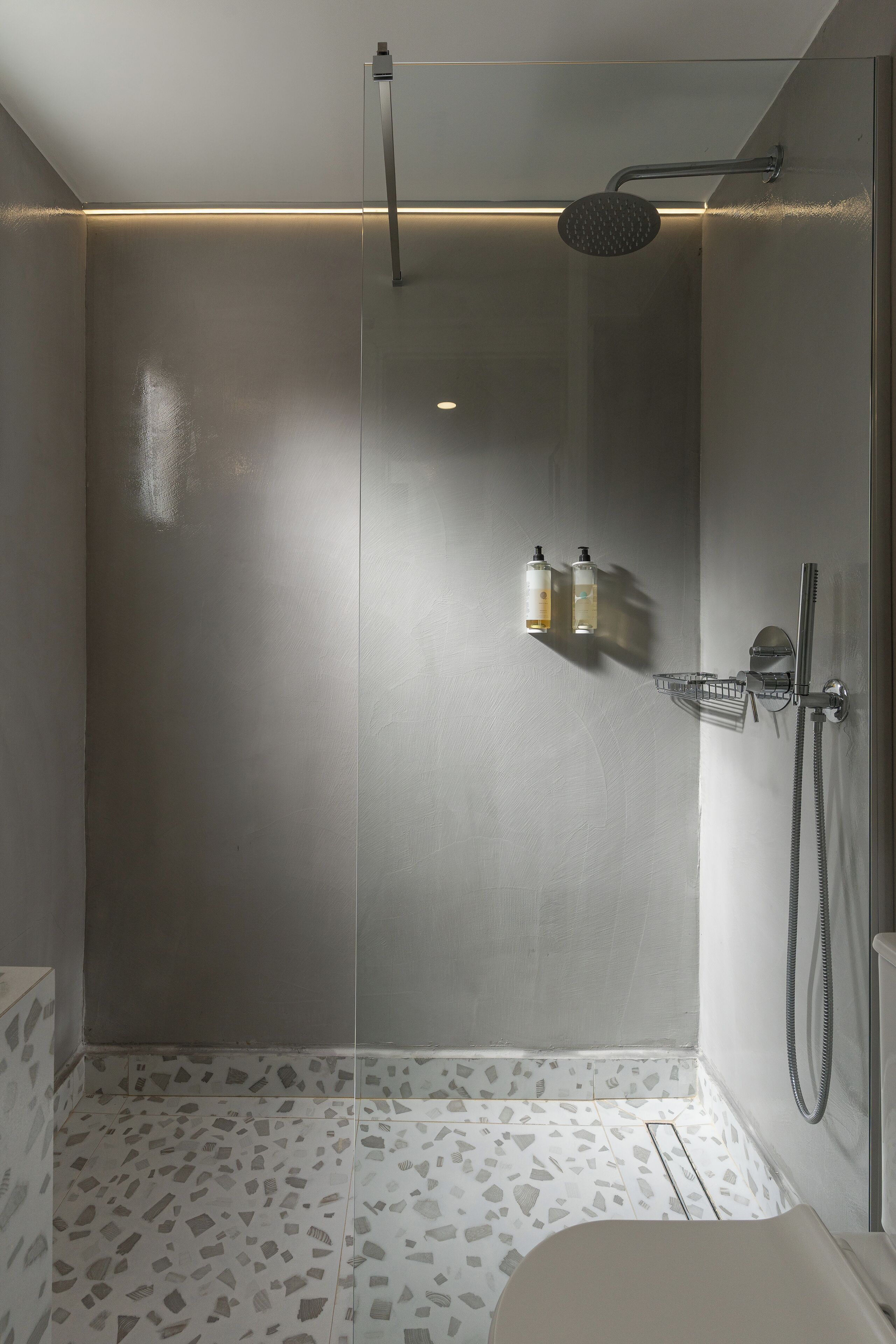 A modern shower with a glass door, a handheld showerhead, and a wall-mounted faucet. There are two bottles of shampoo and conditioner on a shelf.