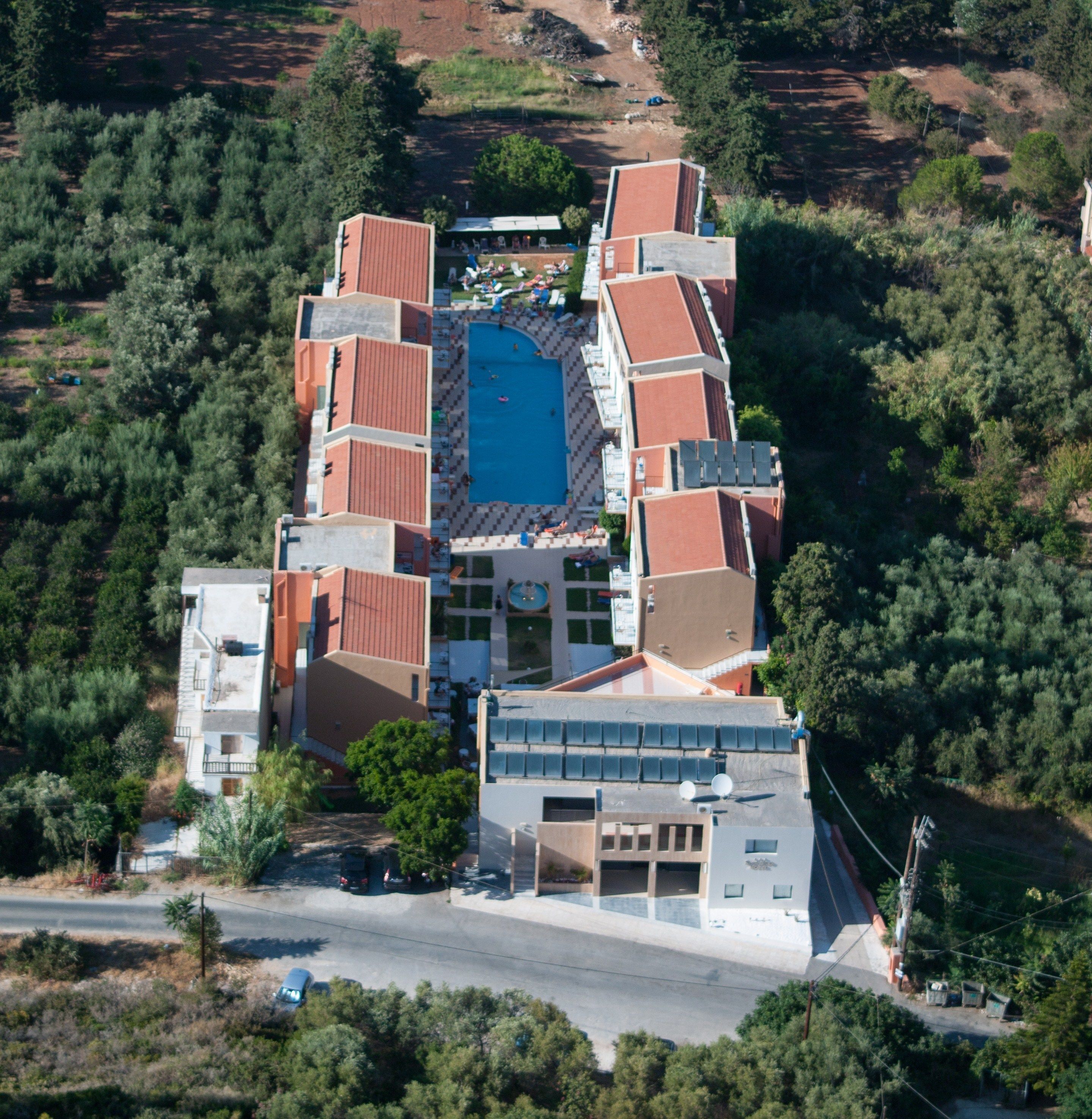 An aerial view of a resort with a central swimming pool, multiple buildings with red roofs, and surrounding greenery.