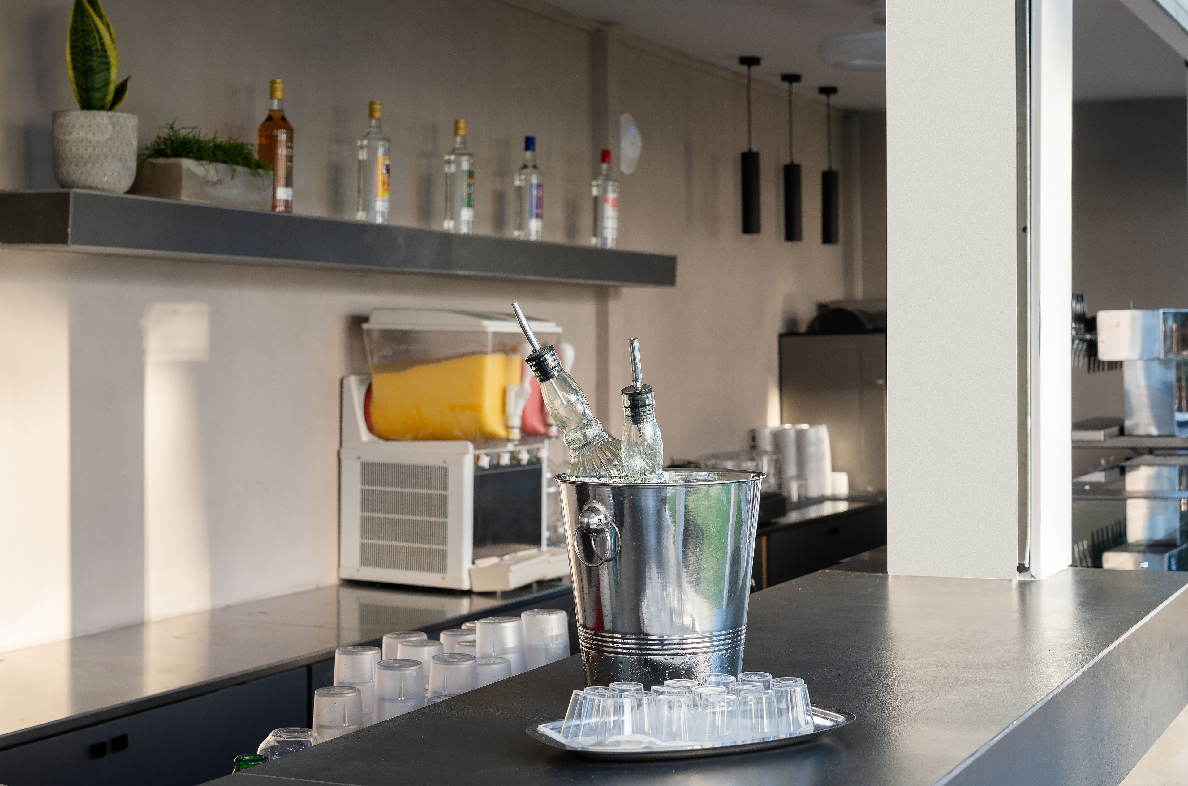 Eine Barbereich mit einem Metalleimer, der Cocktailgläser und Flaschen enthält, einem weißen Kühlschrank und Regalen mit verschiedenen Flaschen.