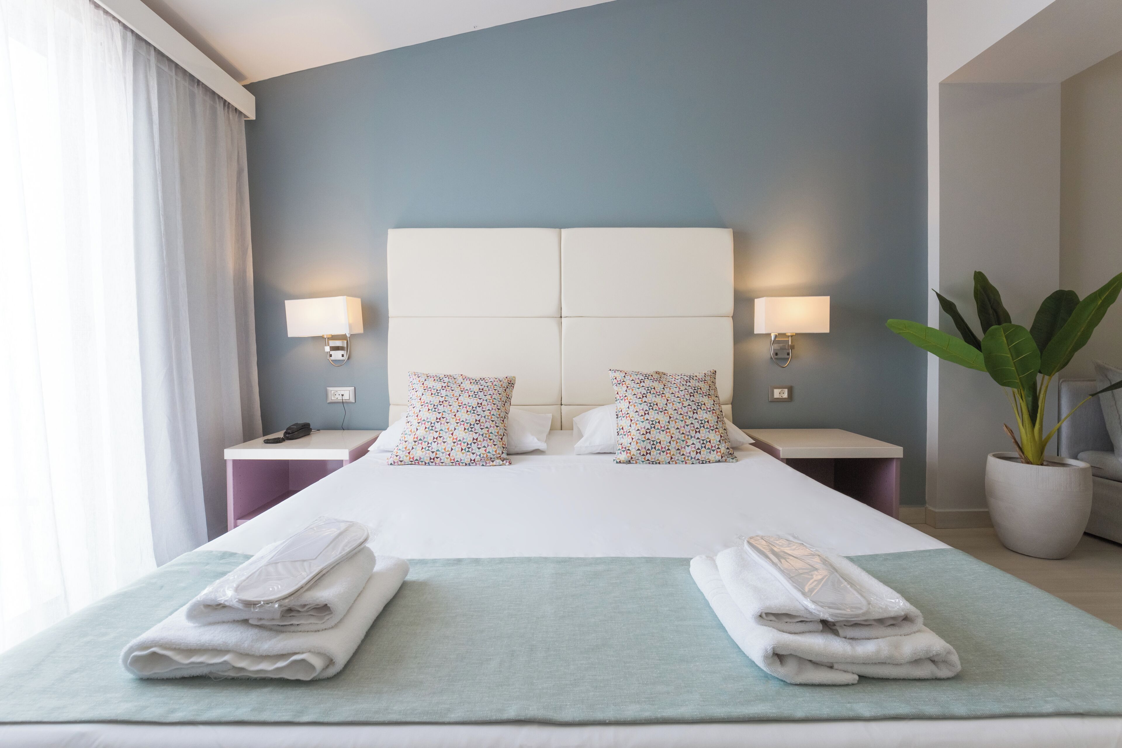 A hotel room with a bed, two bedside tables, a nightstand, a potted plant, and a window with curtains.