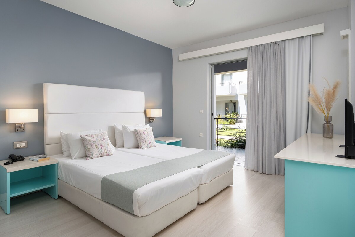Moderni hotellihuone, jossa on suuri sänky, kaksi yöpöytää, työpöytä ja parveke, jossa on verhot.