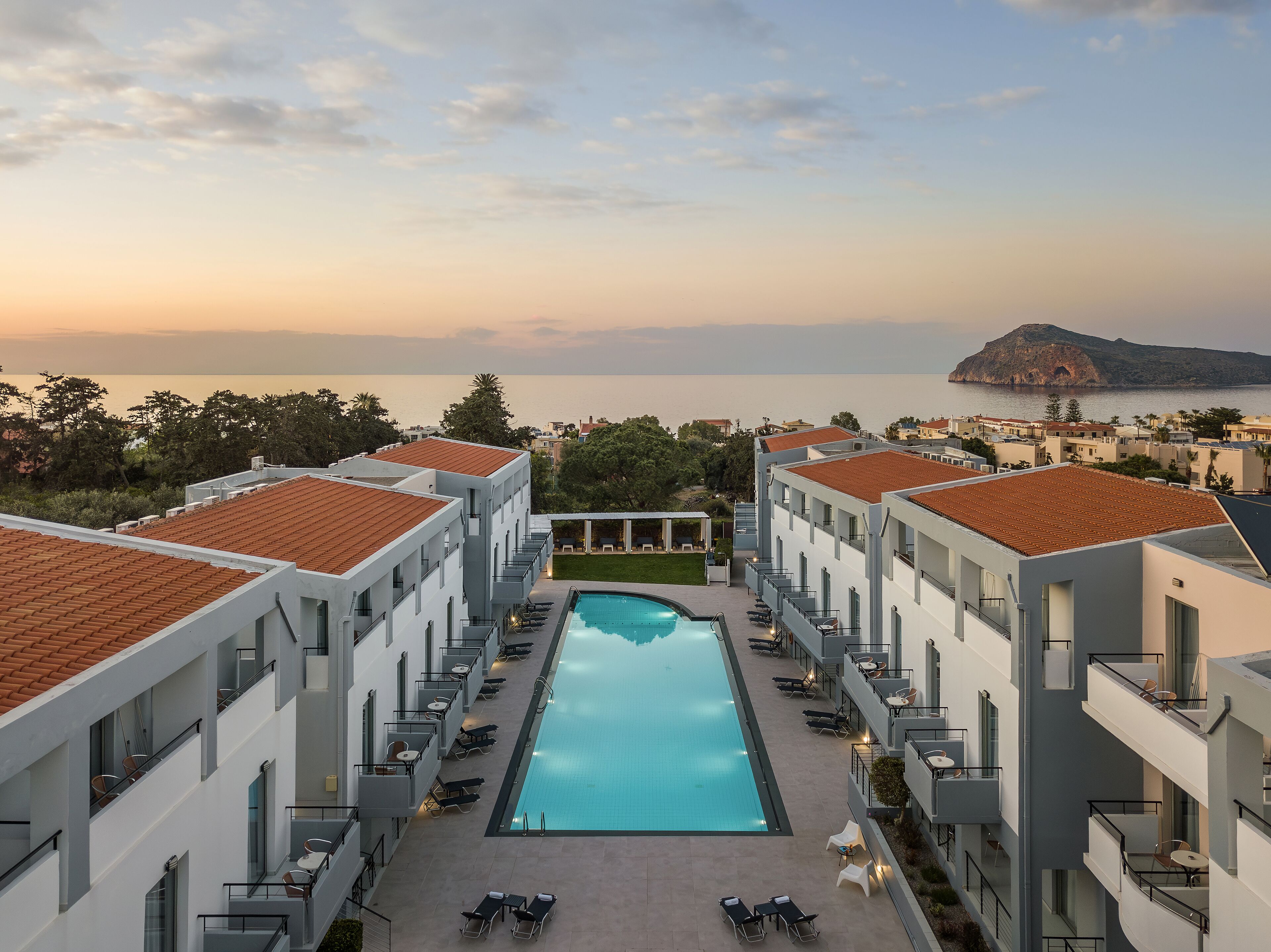 Un hotel moderno con piscina centrale, numerosi balconi e vista sul mare e su una montagna.