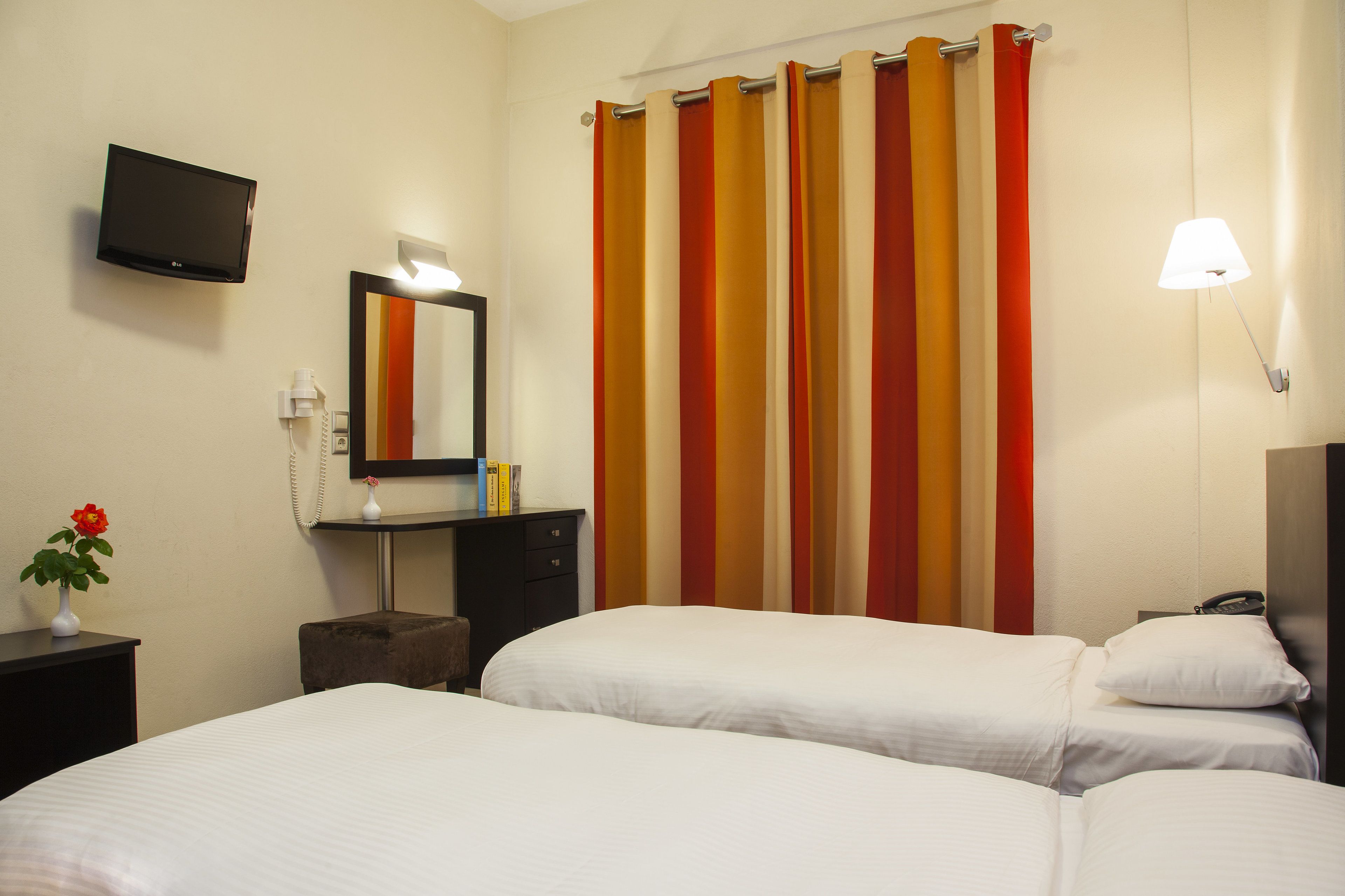 Habitación de hotel con dos camas, un televisor, un espejo, una mesita de noche con un florero y una lámpara.