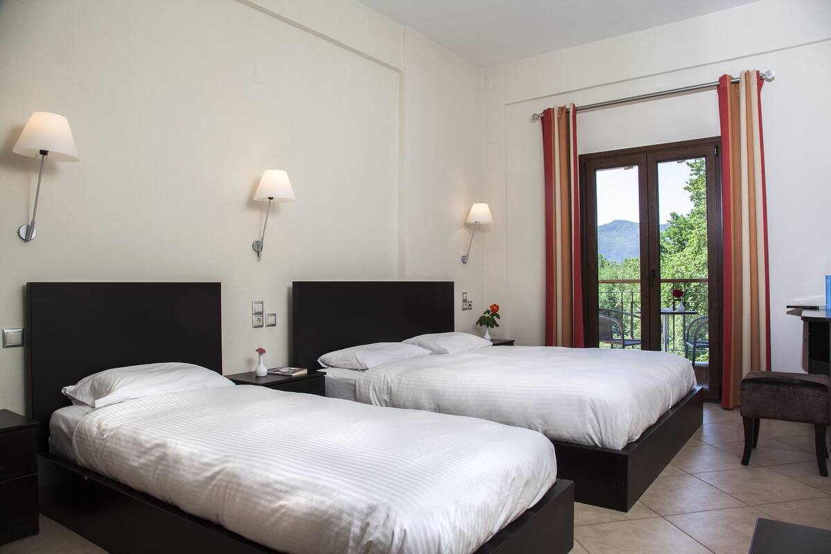 A hotel room with two beds, a desk, and a view of greenery and mountains through the window.