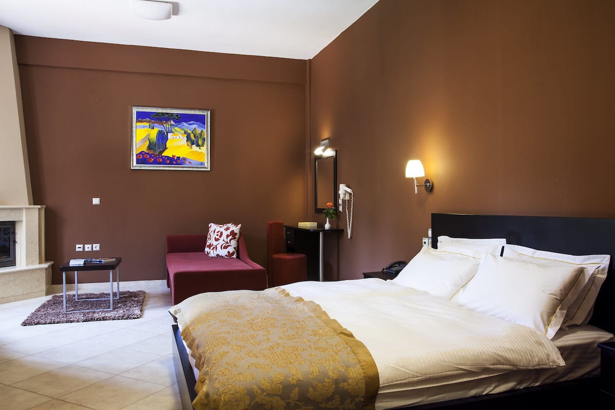 A hotel room with a bed, a desk, a chair, a painting on the wall, and a nightstand with a lamp.