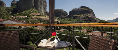 Ein Balkon mit Korbmöbeln, einem Tisch mit offenem Buch und einer Vase mit einer roten Blume, mit Blick auf eine gebirgige Landschaft.