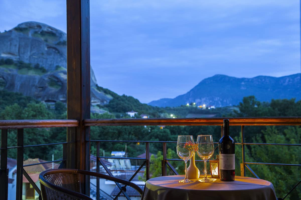 A table with a bottle of wine, two glasses, and a vase with flowers, set against a scenic mountain view from a balcony.