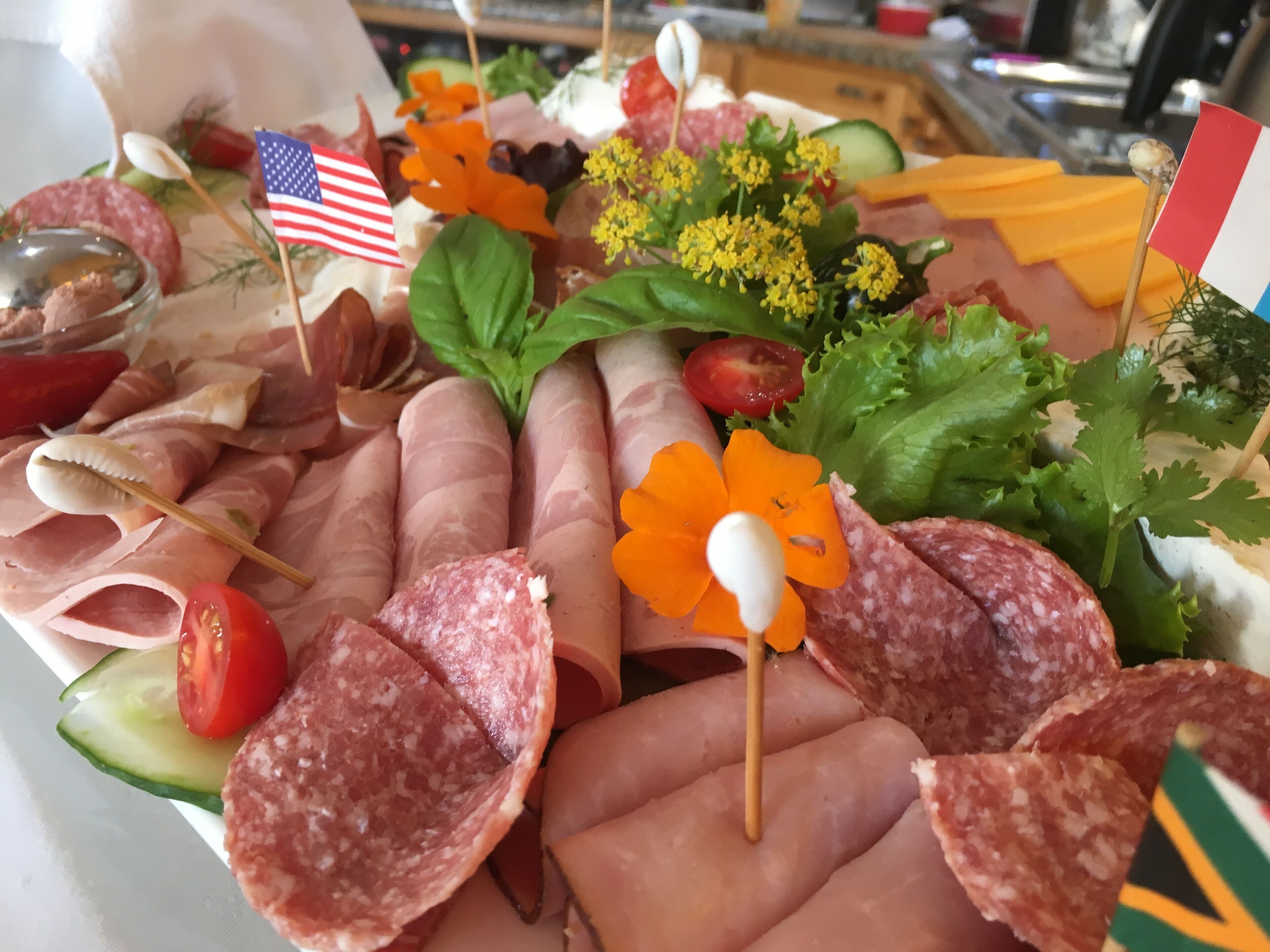 Una selección de embutidos y quesos dispuestos en una bandeja, decorada con flores y pequeñas banderas.