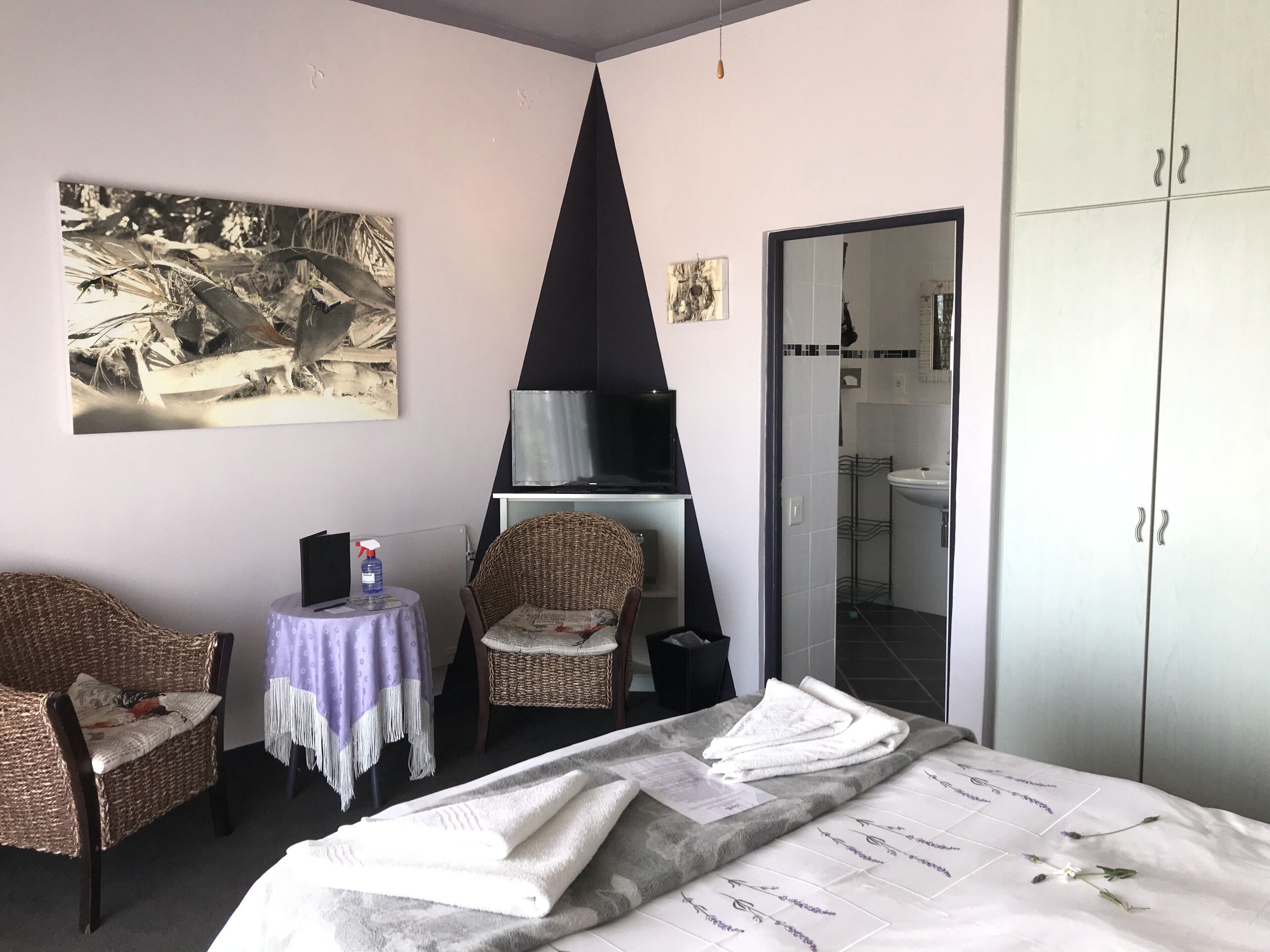 A hotel room with a bed, two wicker chairs, a small table with a spray bottle, a television, and a painting on the wall.