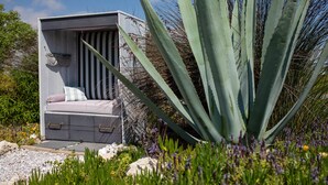 Un espacio al aire libre pequeño y cerrado con un banco acolchado y un armario, rodeado de diversas plantas y una gran planta de agave.