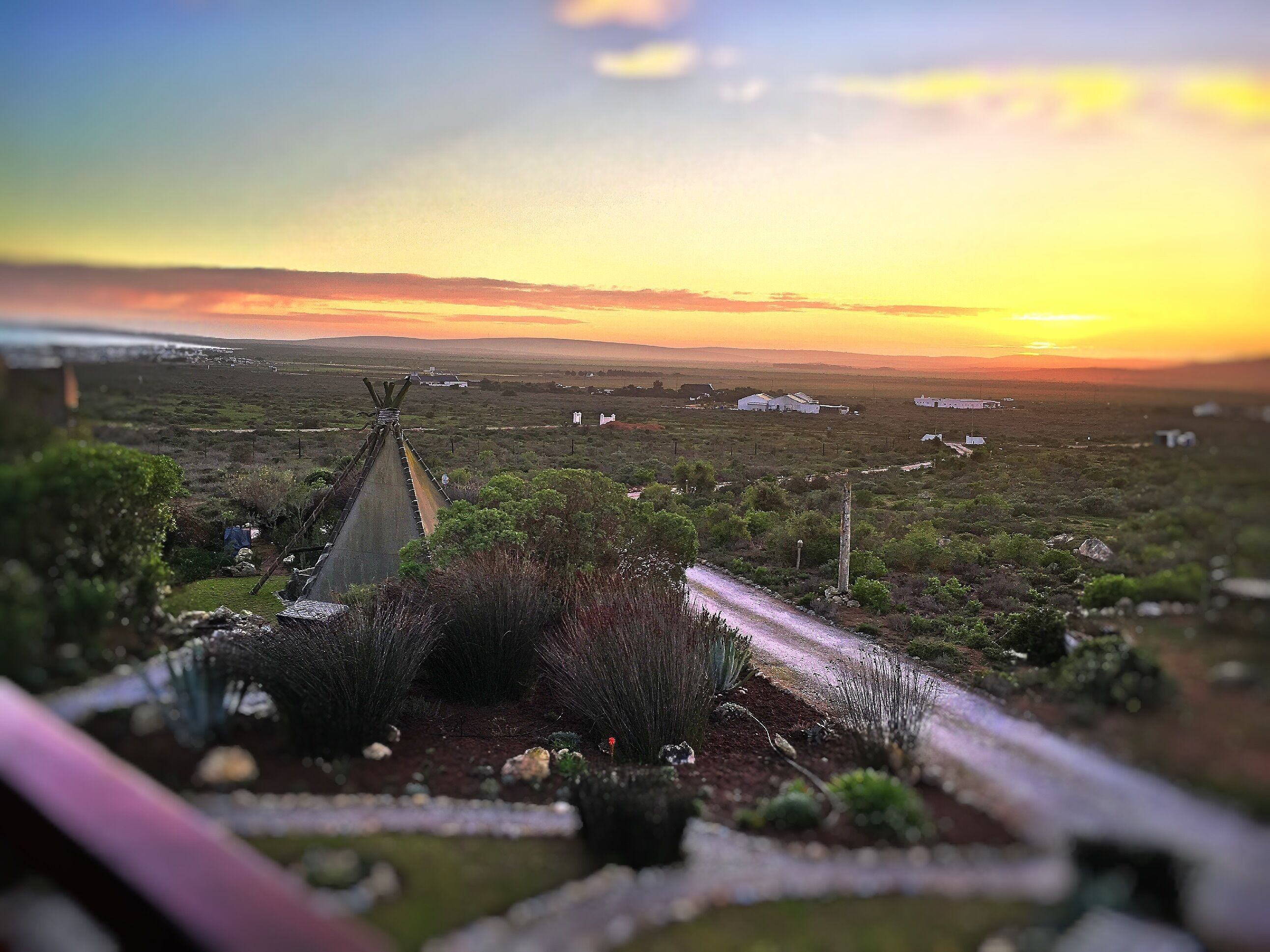 Landelijke omgeving met heldere lucht en een zonsondergang. Er zijn velden, enkele verspreide gebouwen en een weg die in de verte leidt.