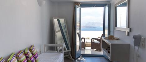 A bedroom with a bed, pillows, a bedside table, a mirror, and a view of the sea through an open door.