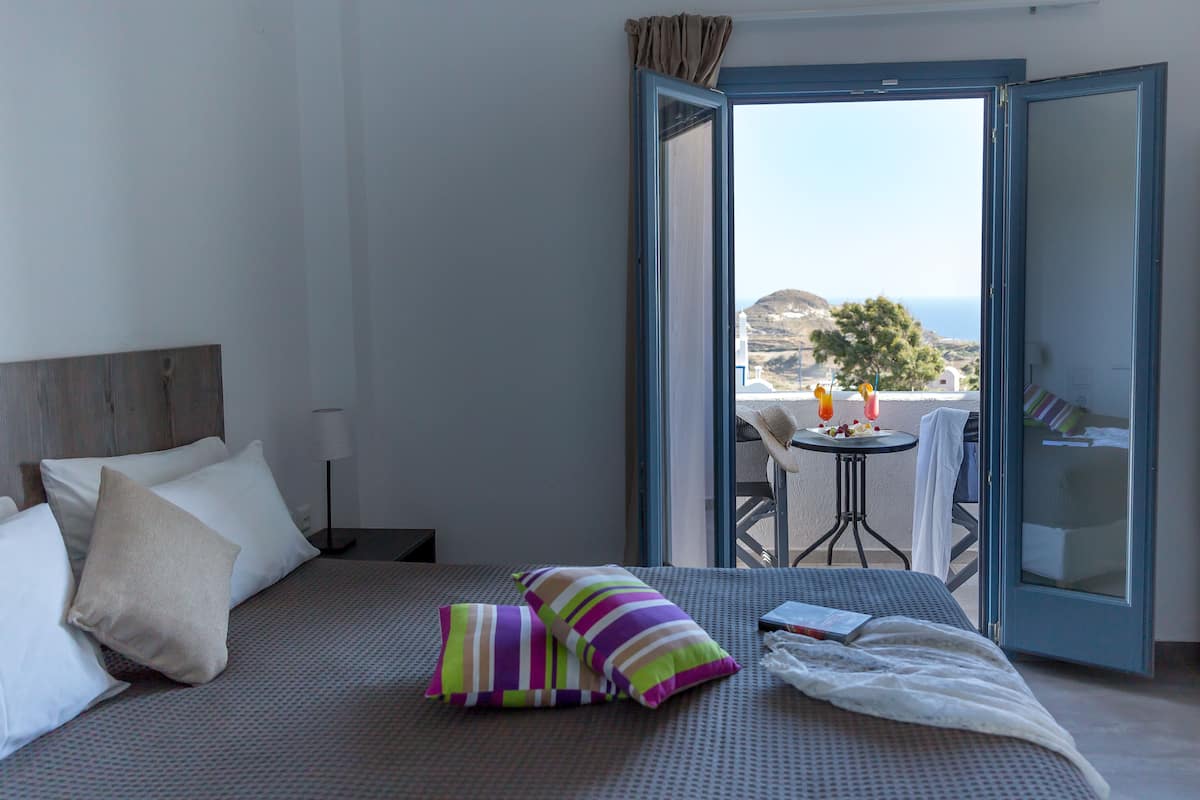 A hotel room with a bed, pillows, a bedside table, a lamp, and a view of the sea through an open door.