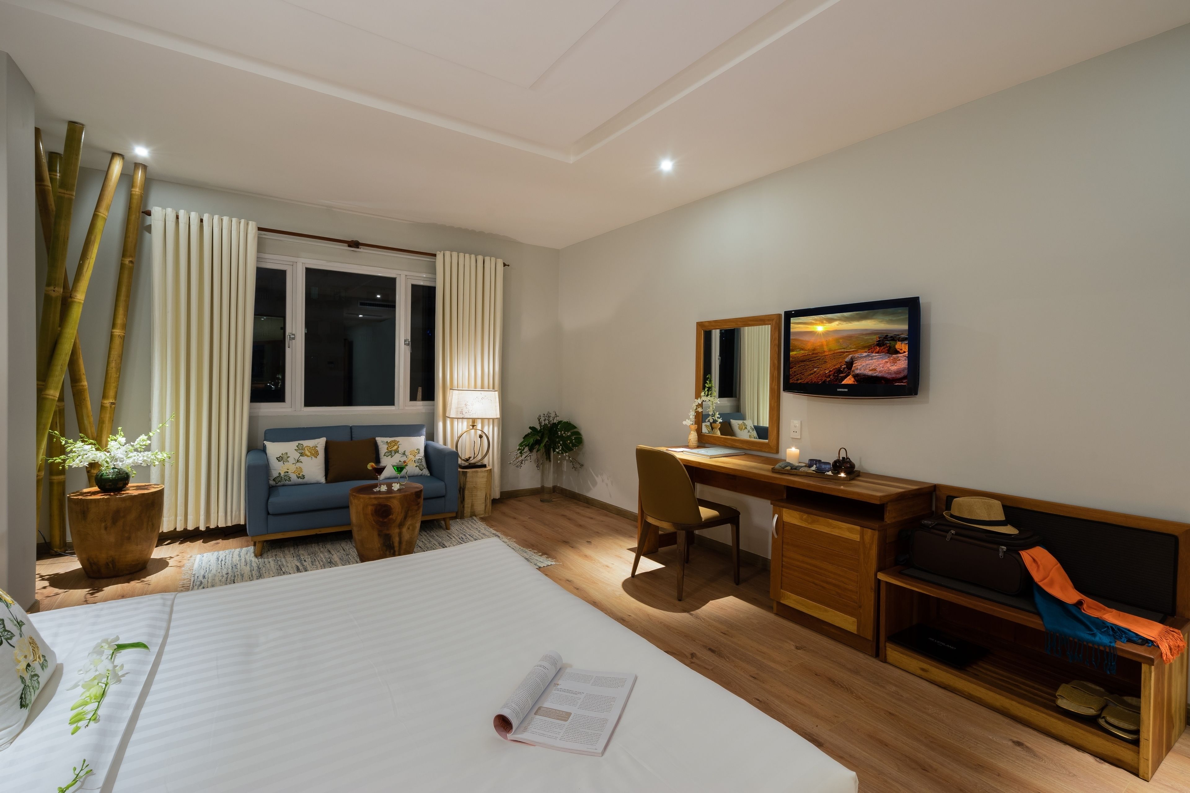 A hotel room with a large bed, a desk, a chair, a sofa, a wooden bench, a mirror, and a television.