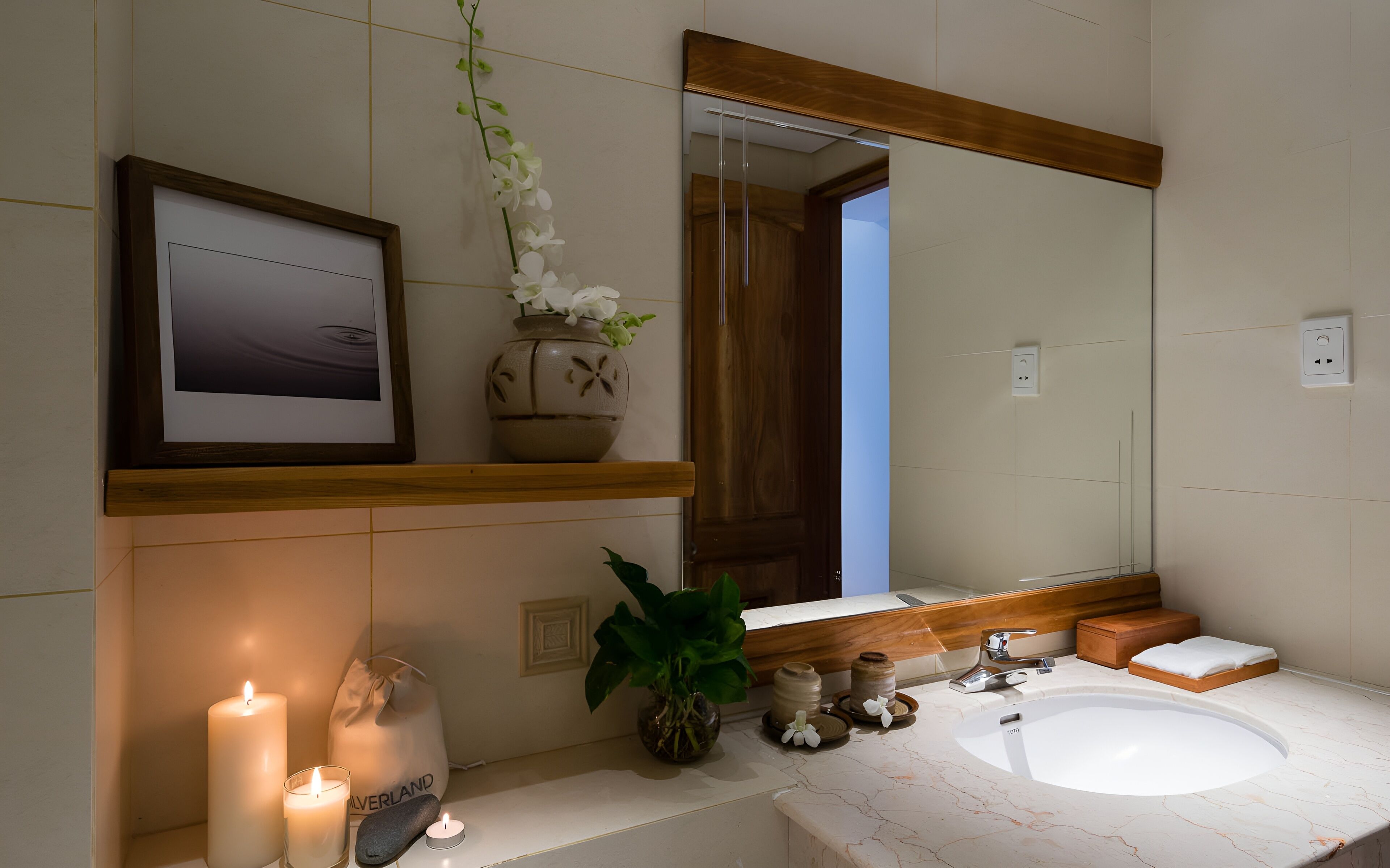 Un baño con un espejo grande, un lavabo, un cuadro enmarco, un florero con flores y velas.