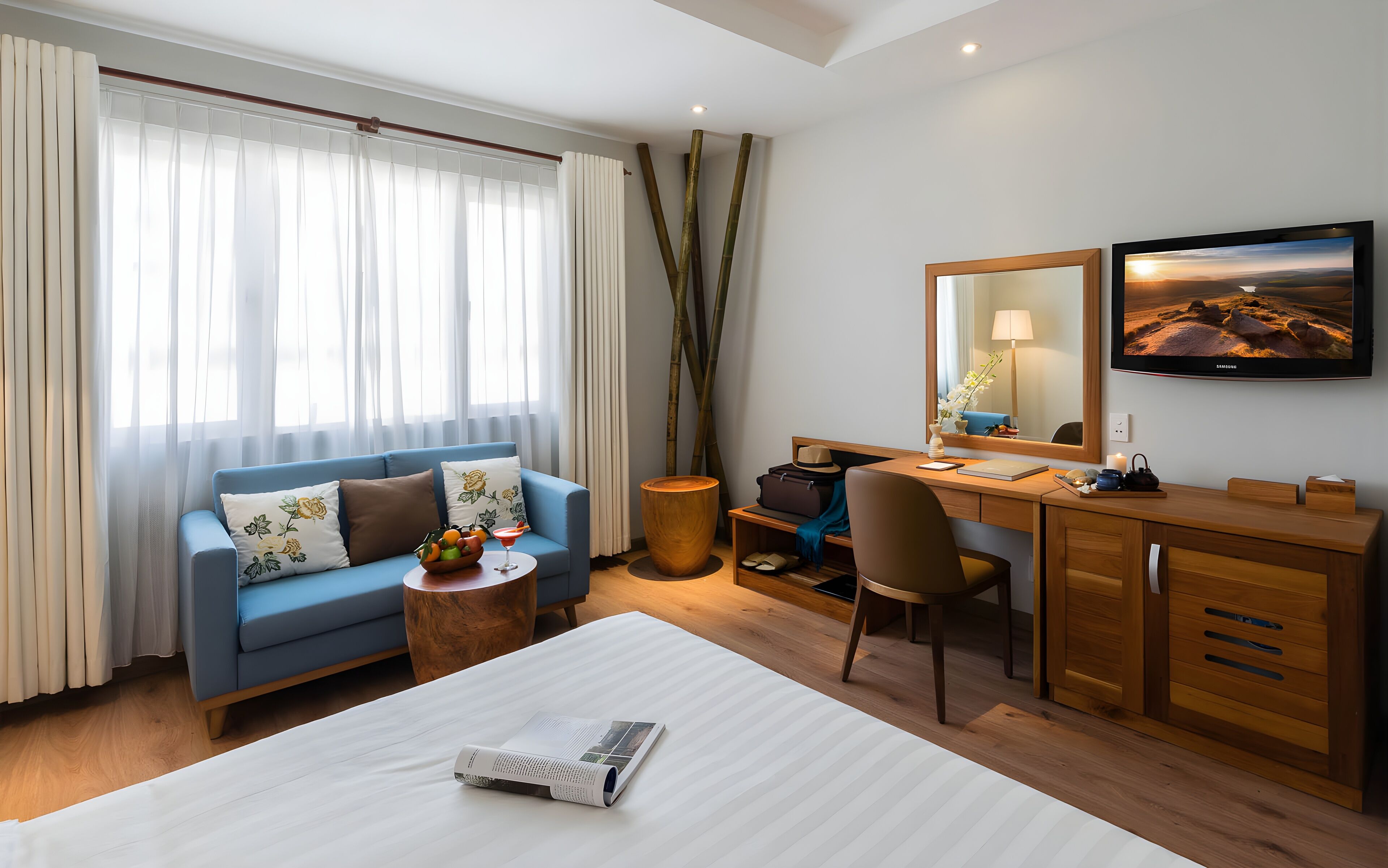 A hotel room with a blue sofa, a wooden desk, a bed with a white comforter, and a large window with sheer curtains.