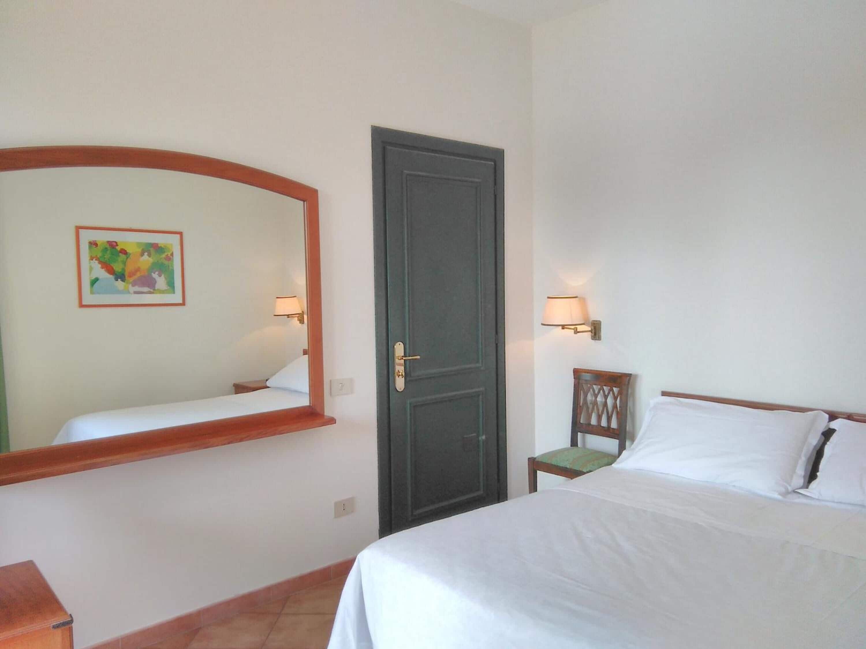 Een hotelkamer met een bed, een stoel, een bureau, een spiegel, een deur en een wandlamp.