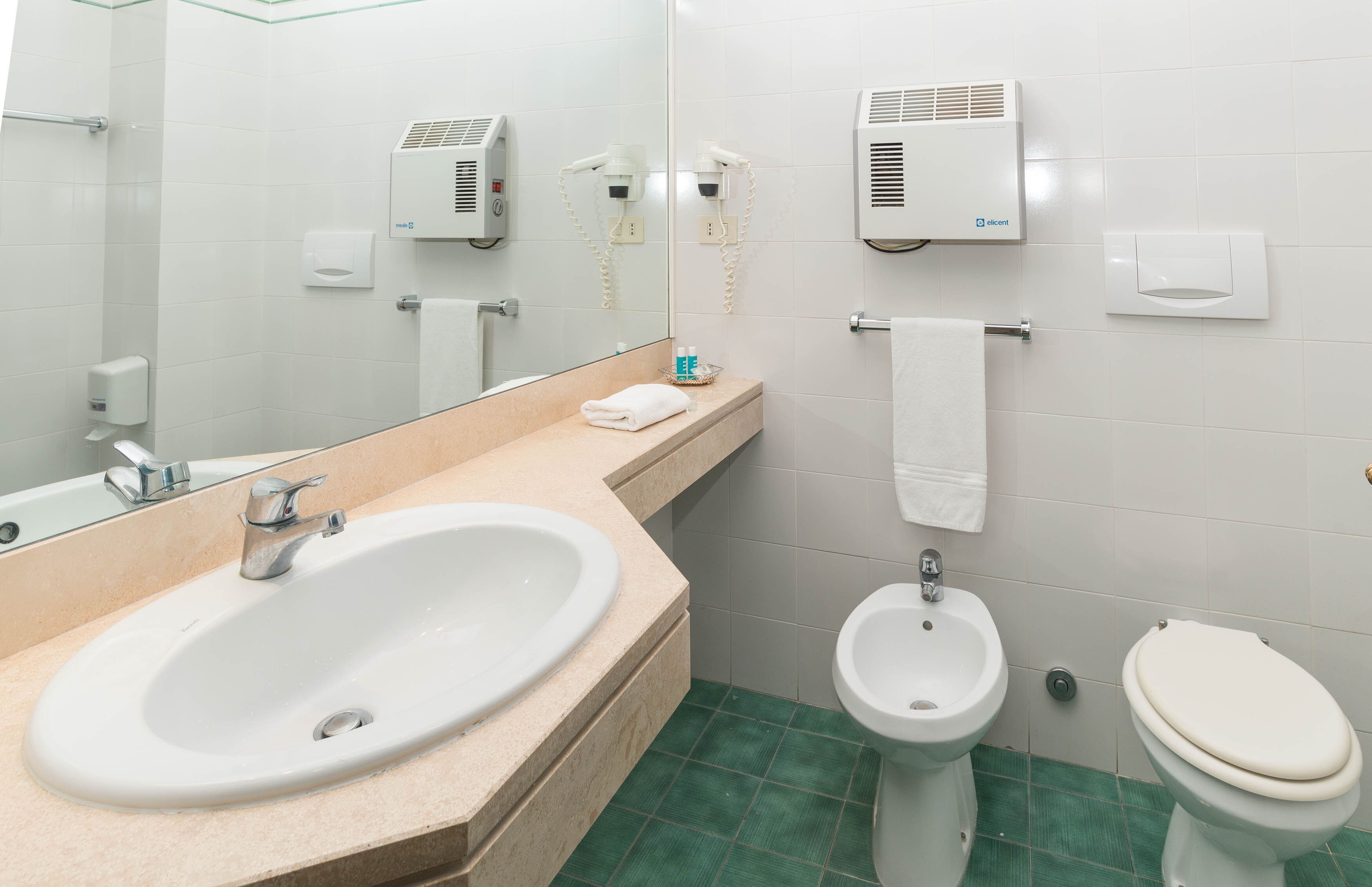 A bathroom with a sink, bidet, toilet, towel rack, and hair dryers mounted on the wall.