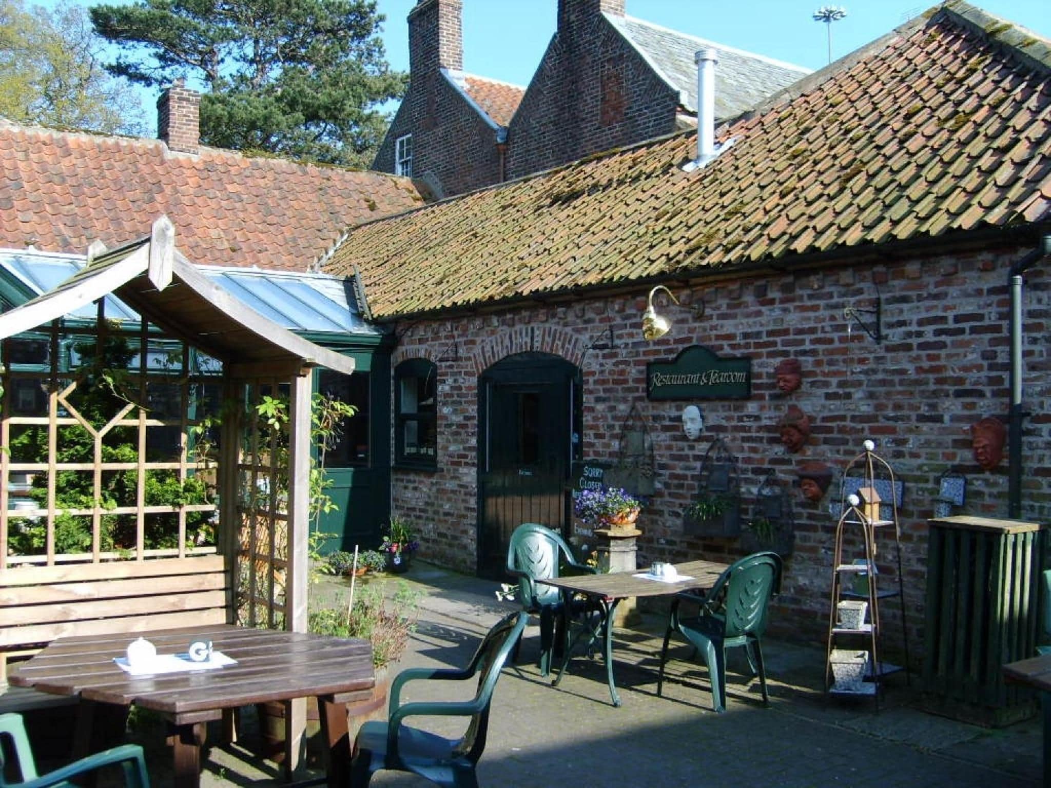 A brick building with a tiled roof, a sign reading 'Restaurant & Bar,' outdoor seating with tables and chairs, and a garden area with plants.