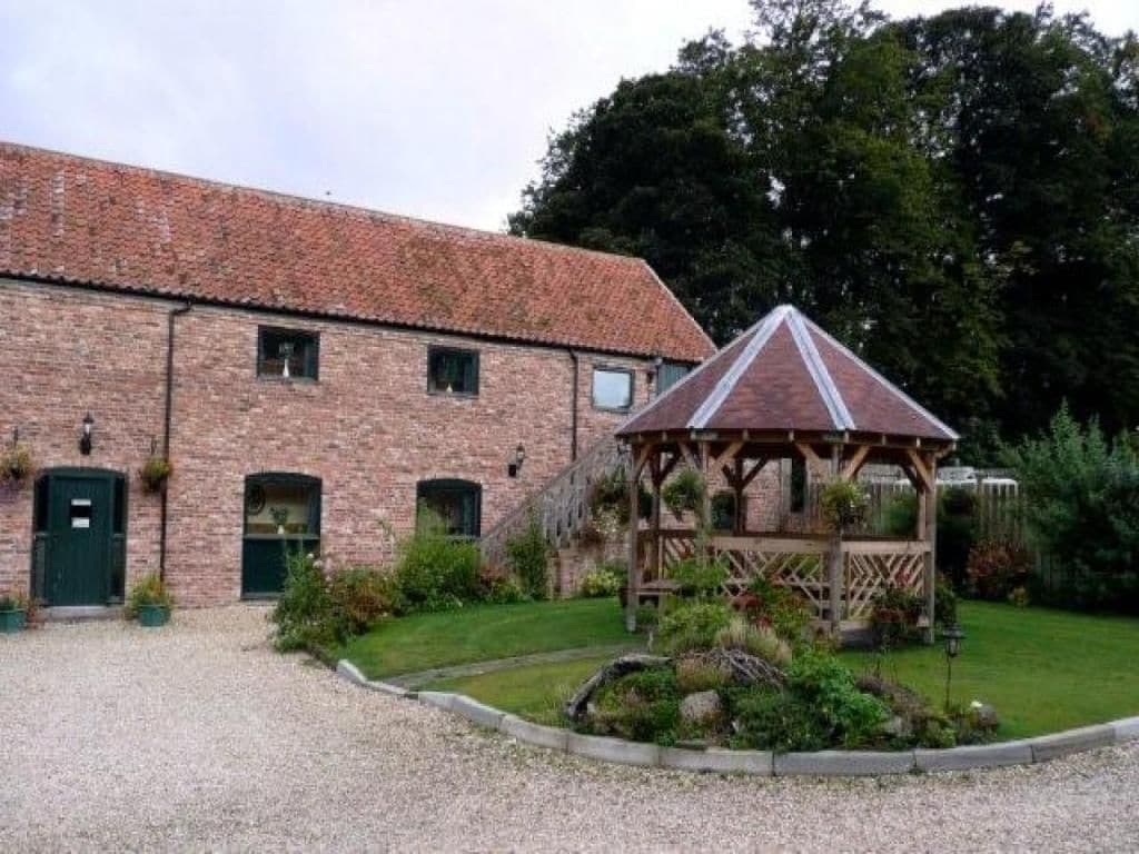 Un bâtiment en briques doté d’un espace extérieur couvert avec des sièges, d’une allée en gravier et d’un jardin bien entretenu.