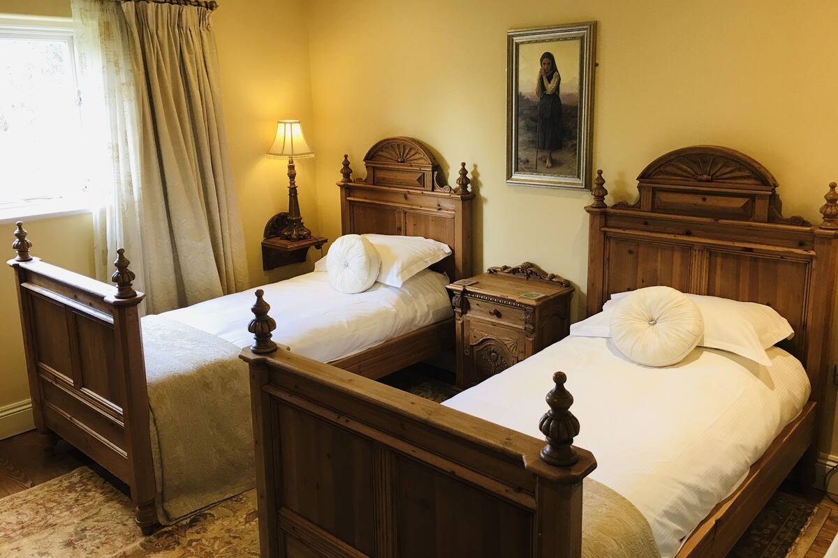 Une chambre avec deux lits en bois, une table de chevet avec une lampe, un tableau au mur et une fenêtre avec des rideaux.